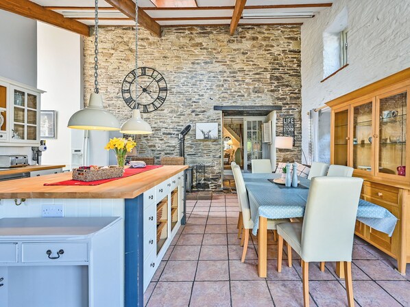 Une cuisine rustique avec un mur en pierre, des poutres en bois, une grande horloge et un coin repas comprenant une table et des chaises.
