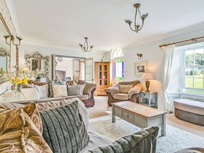 Un salon confortable avec un canapé, un fauteuil et une petite table. On y trouve un miroir, un lustre et une fenêtre avec des rideaux.