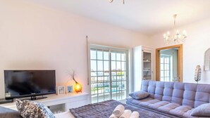 Ein modernes Wohnzimmer mit einem großen Flachbildfernseher, einer Couch, einem Bett mit einer braunen Decke und Blick ins Freie durch ein großes Fenster.