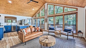 A spacious living area with a wooden ceiling, large windows, a leather sofa, and a round coffee table. - 'Foxtail' Cabin Retreat: Steps to Beaver Lake! (Rogers)