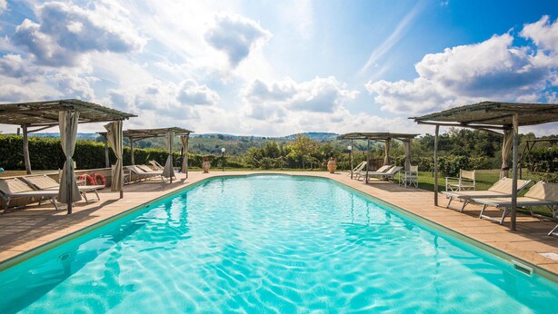 Ein klares Schwimmbecken mit Sonnenschirmen und Liegestühlen, umgeben von einem angelegten Garten und Blick auf ferne Hügel.