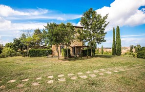 Un bâtiment rustique en pierre, doté d’un patio couvert, entouré d’une pelouse verdoyante et d’arbres.