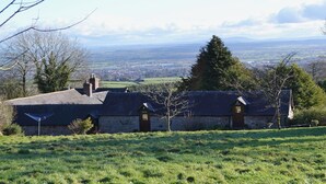 Uma casa de pedra com telhado inclinado, um pequeno jardim e vista para uma cidade distante e colinas.