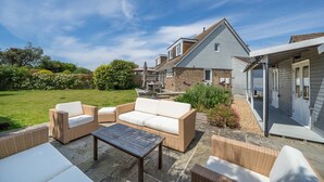 En terrasse med fletmøbler og et stentabel, et hus med et blåt tag, og en have med forskellige planter.