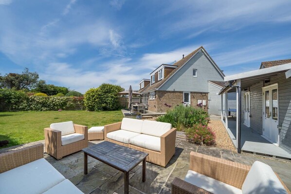 Un espace patio aménagé avec des meubles en osier et une table en pierre, une maison avec un toit bleu et un jardin où poussent différentes espèces de plantes.