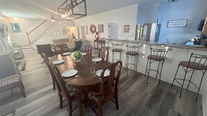 Un comedor amplio con una larga mesa de madera dispuesta para seis comensales, rodeada de sillas. Se aprecia una cocina con barra de fondo.
