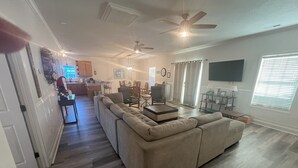 Un salón amplio con un sofá seccional beige grande, una zona de comedor con mesa y sillas, una cocina al fondo y un ventilador de techo.
