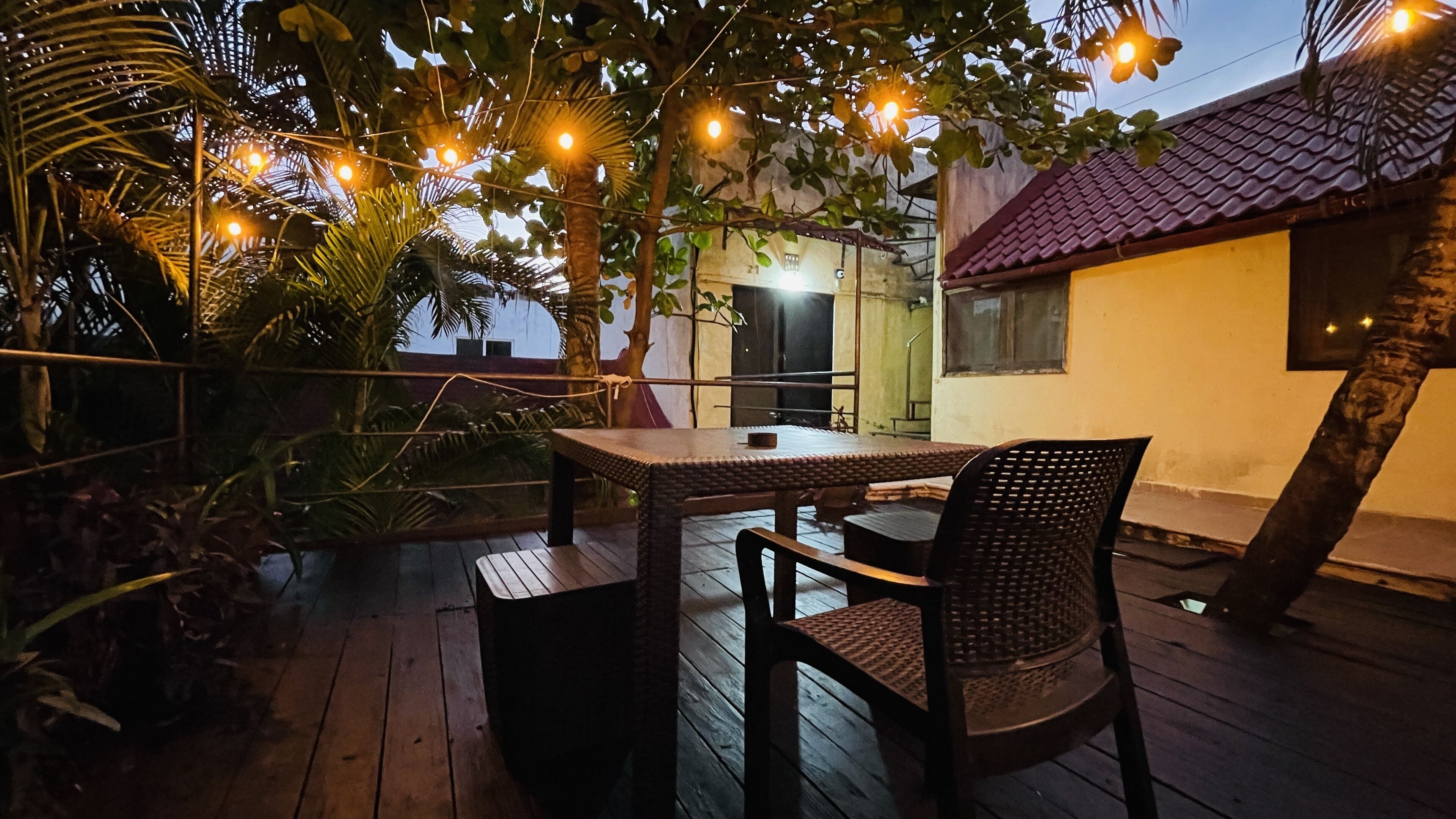 An outdoor patio with a table and chairs, surrounded by plants and string lights.