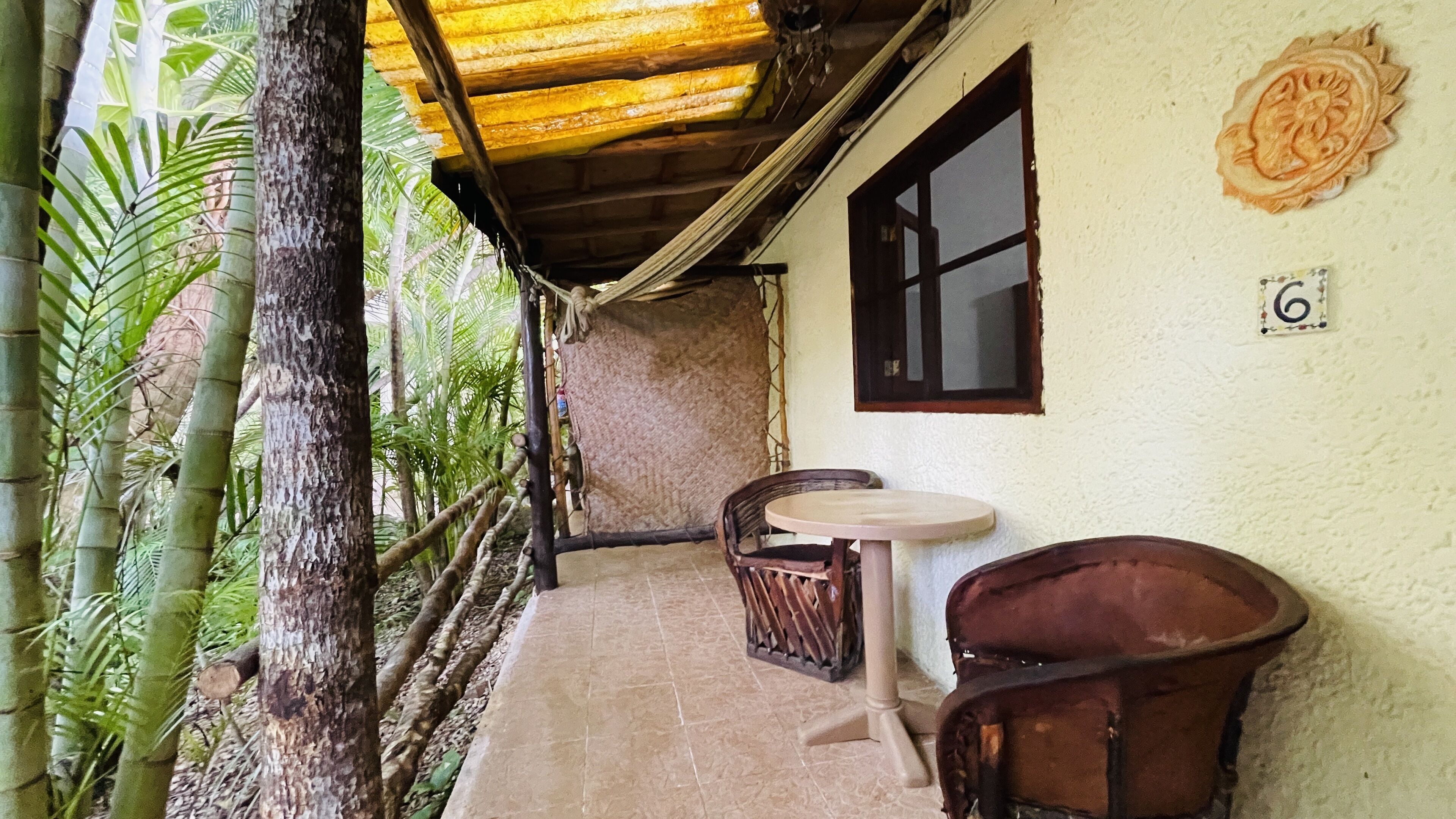 A covered outdoor seating area with a table and chairs, surrounded by bamboo and palm trees.