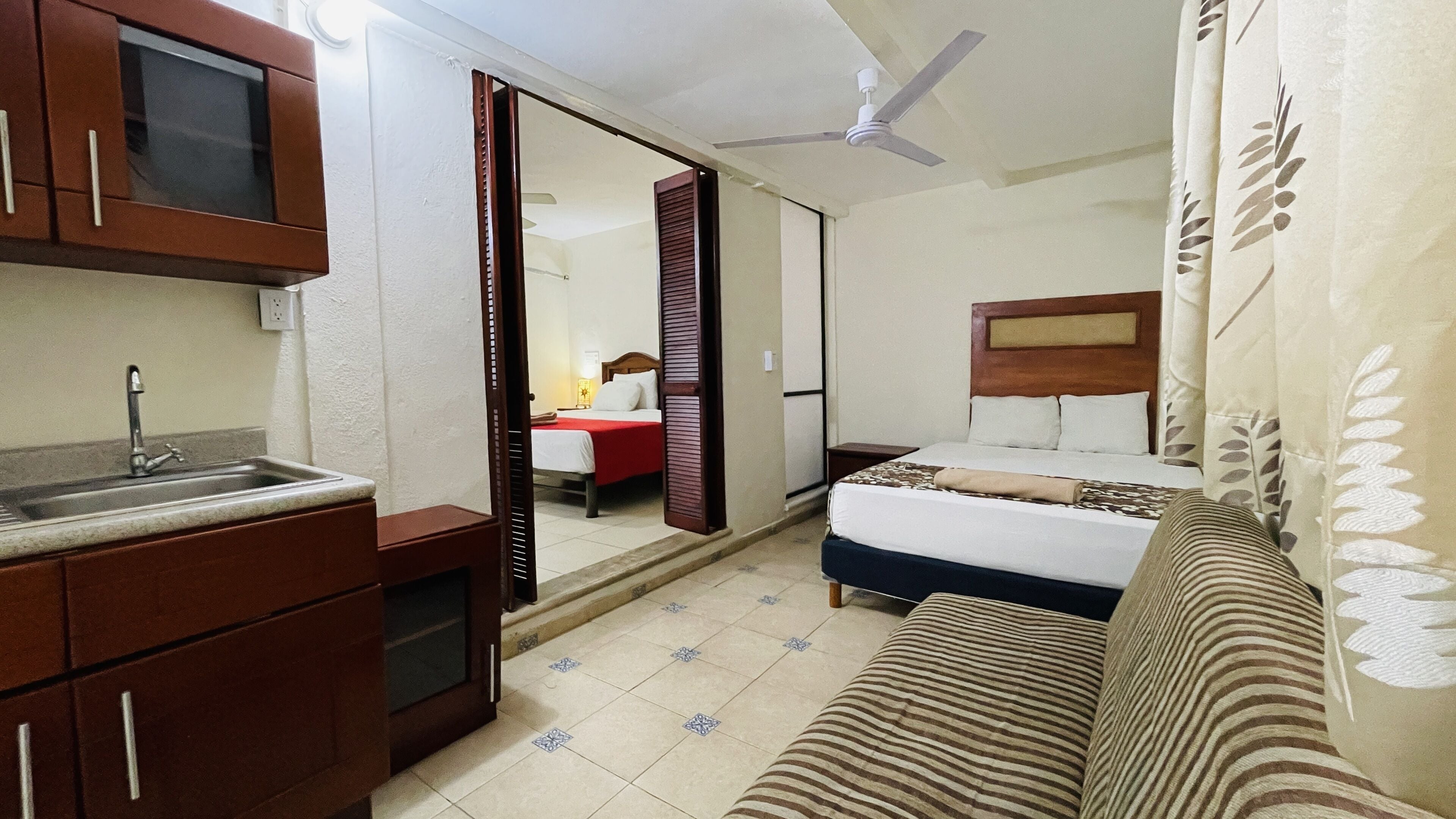 Una habitación de hotel compacta con kitchenette, una cama con cabecera, un sofá y un ventilador de techo.