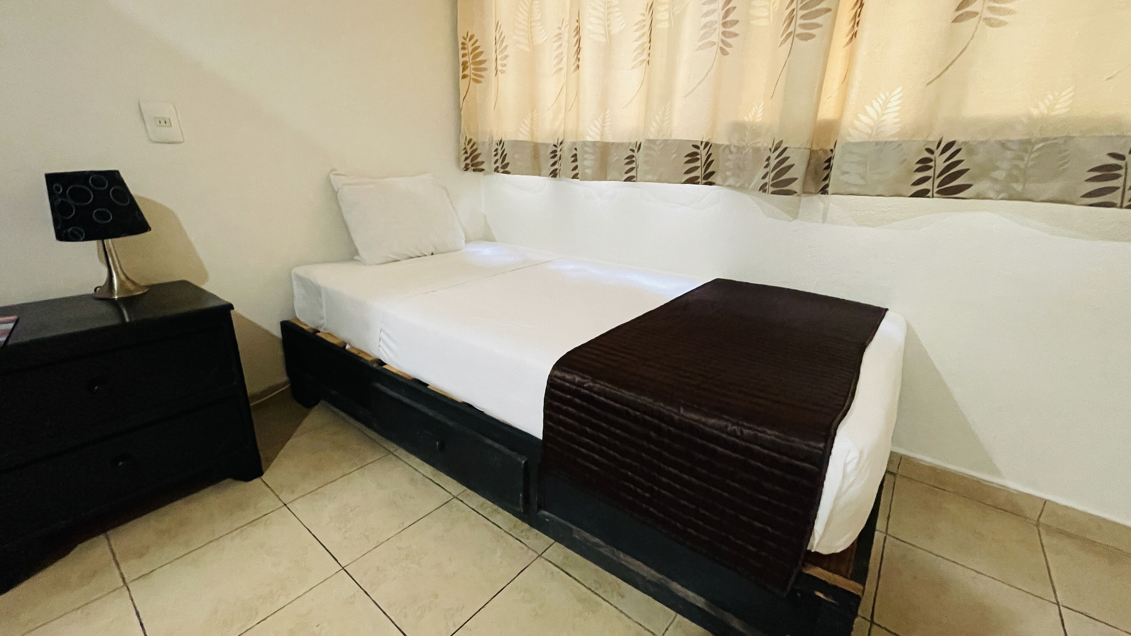 A single bed with a white mattress and a brown blanket, a black nightstand with a lamp, and a power outlet on the wall.