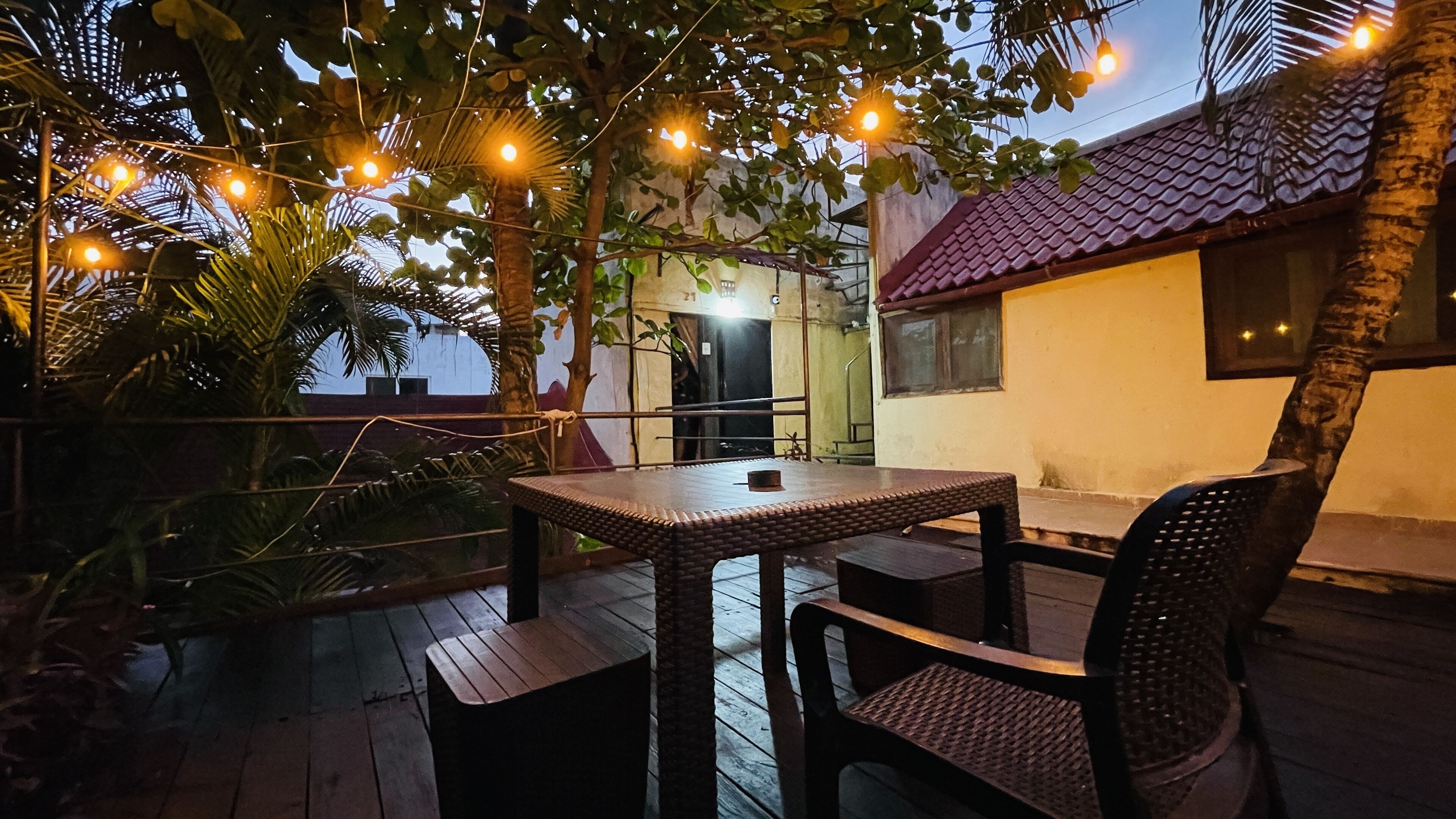 Un patio exterior con una mesa y sillas, rodeado de plantas y luces colgantes.