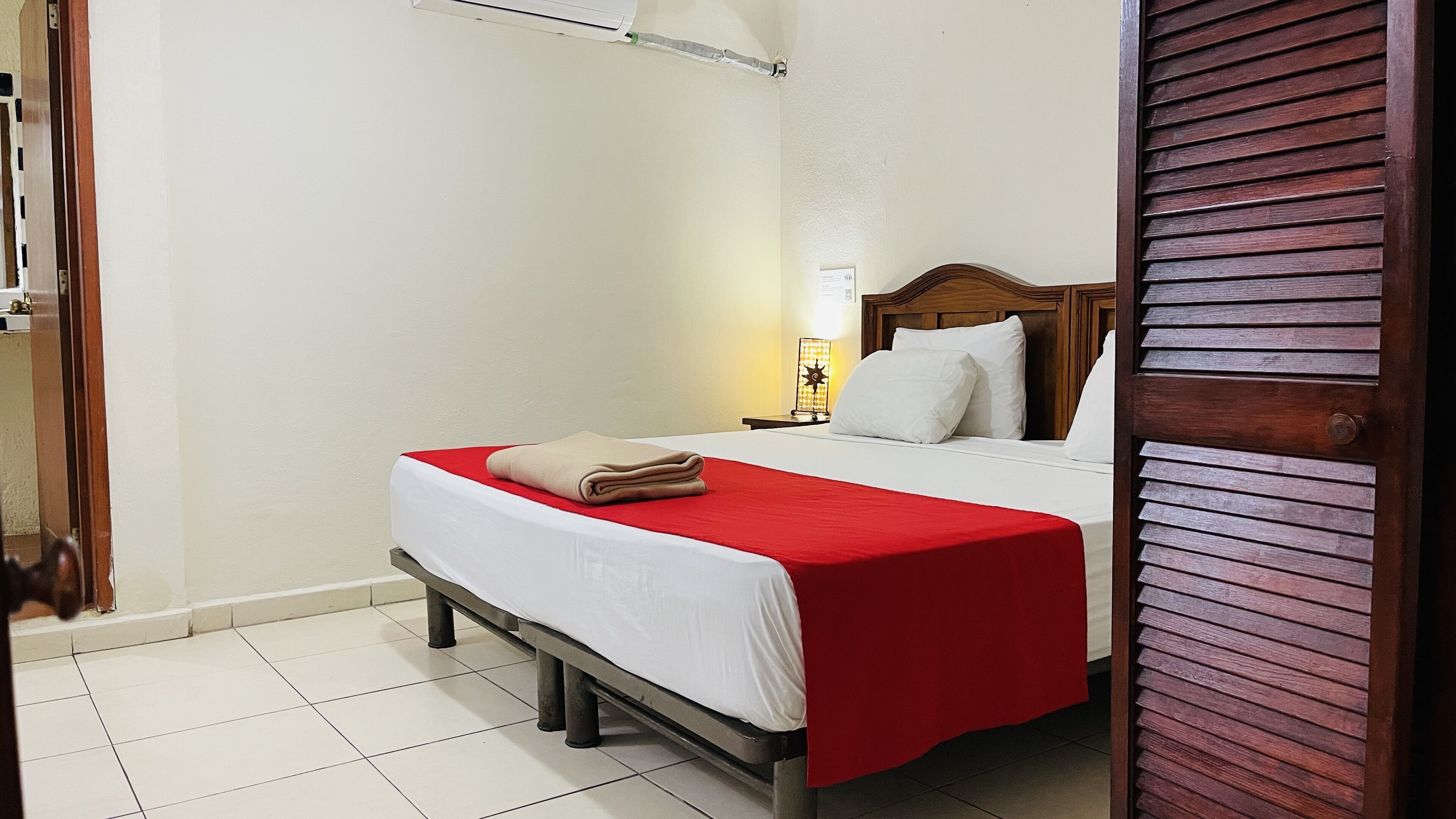 A neatly made bed with a red blanket and a folded beige towel, a wooden headboard, a bedside lamp, and a white wall with a window.