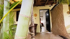 Estructura de bambú con un tapete tejido, una puerta y piso de baldosas que conducen a una habitación con cama y mesita de noche.