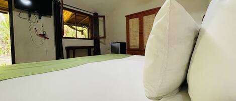 A room with a bed, a television mounted on the wall, a window with a view of greenery, and a door with a traditional design.