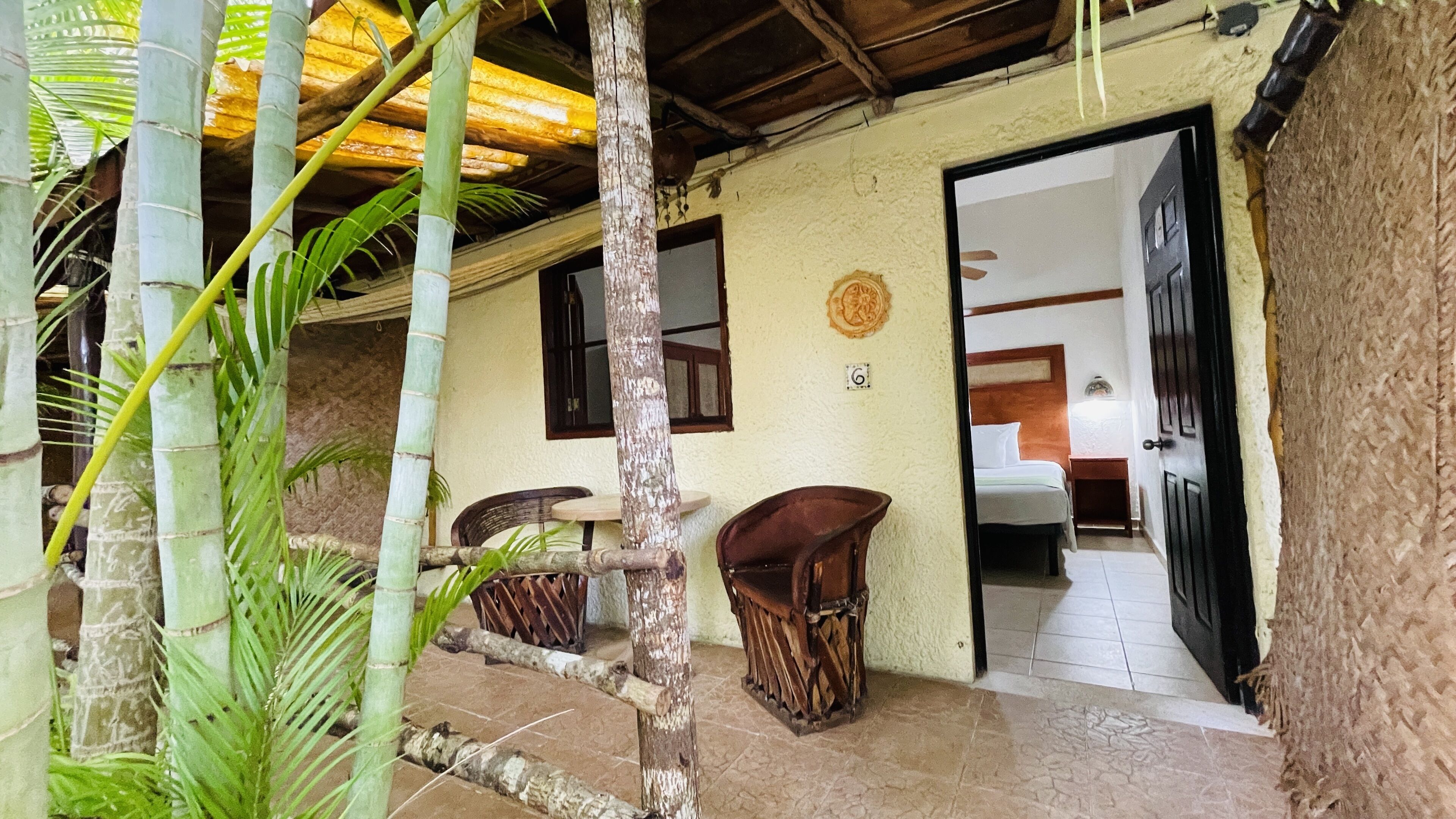 A rustic building with a tiled patio, bamboo plants, and a wooden door leading to a bedroom with a bed and nightstand.