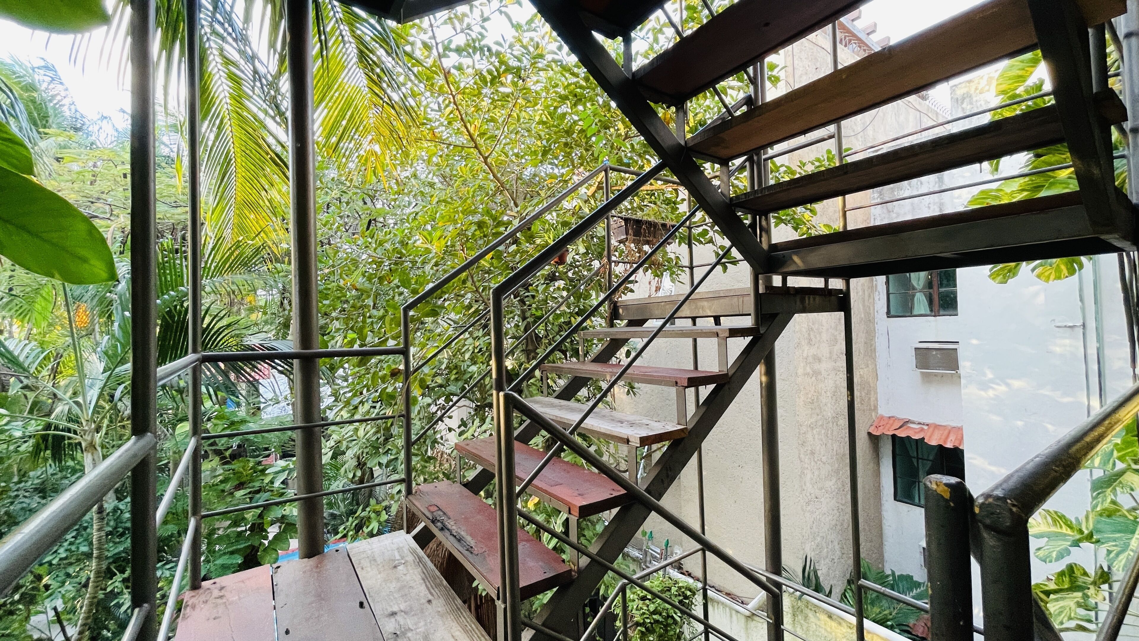 A staircase with metal railings and wooden steps, surrounded by lush greenery and a building in the background.