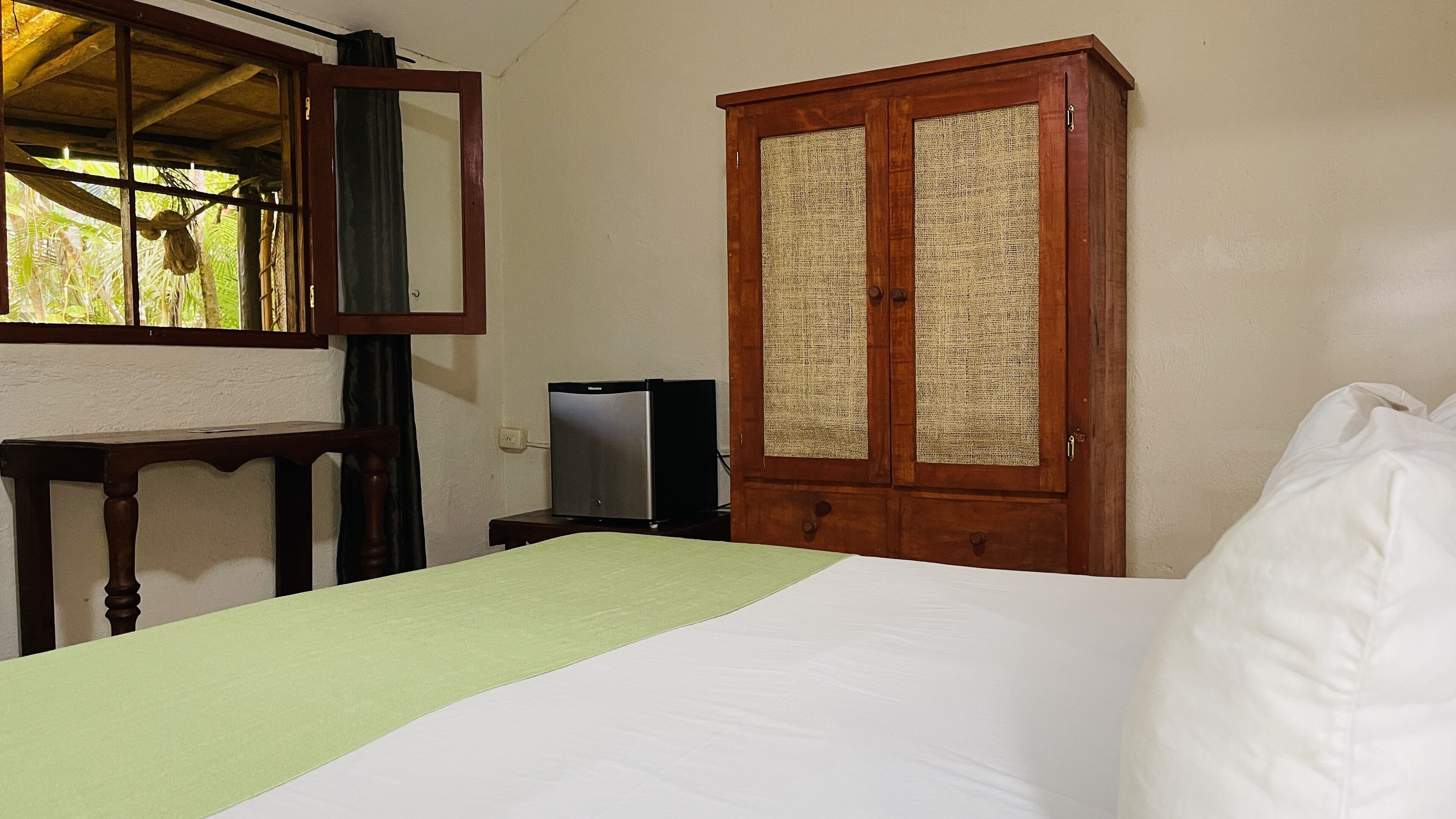 A bedroom with a bed, a wooden wardrobe with a woven door, a small table with a television on it, and a window with a view of greenery.