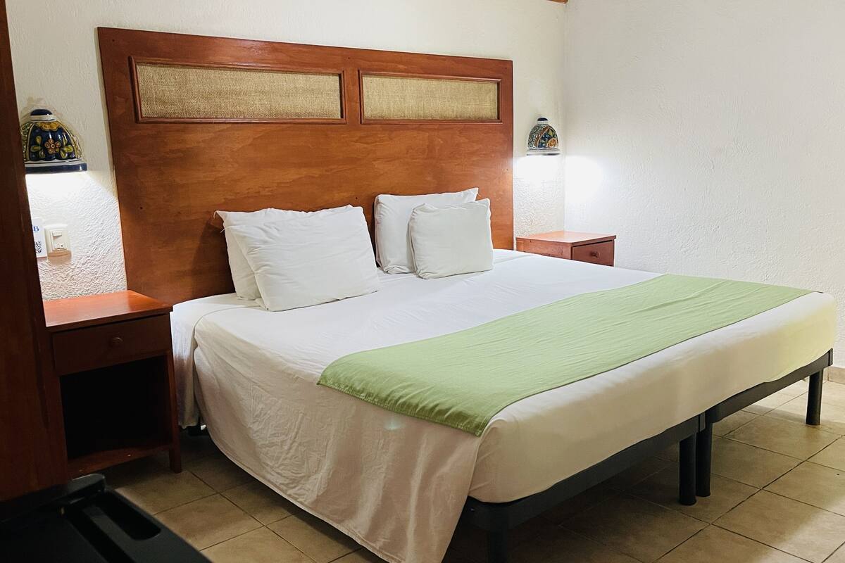A neatly made bed with white linens and a green throw, a wooden headboard, bedside tables with lamps, and a tiled floor.