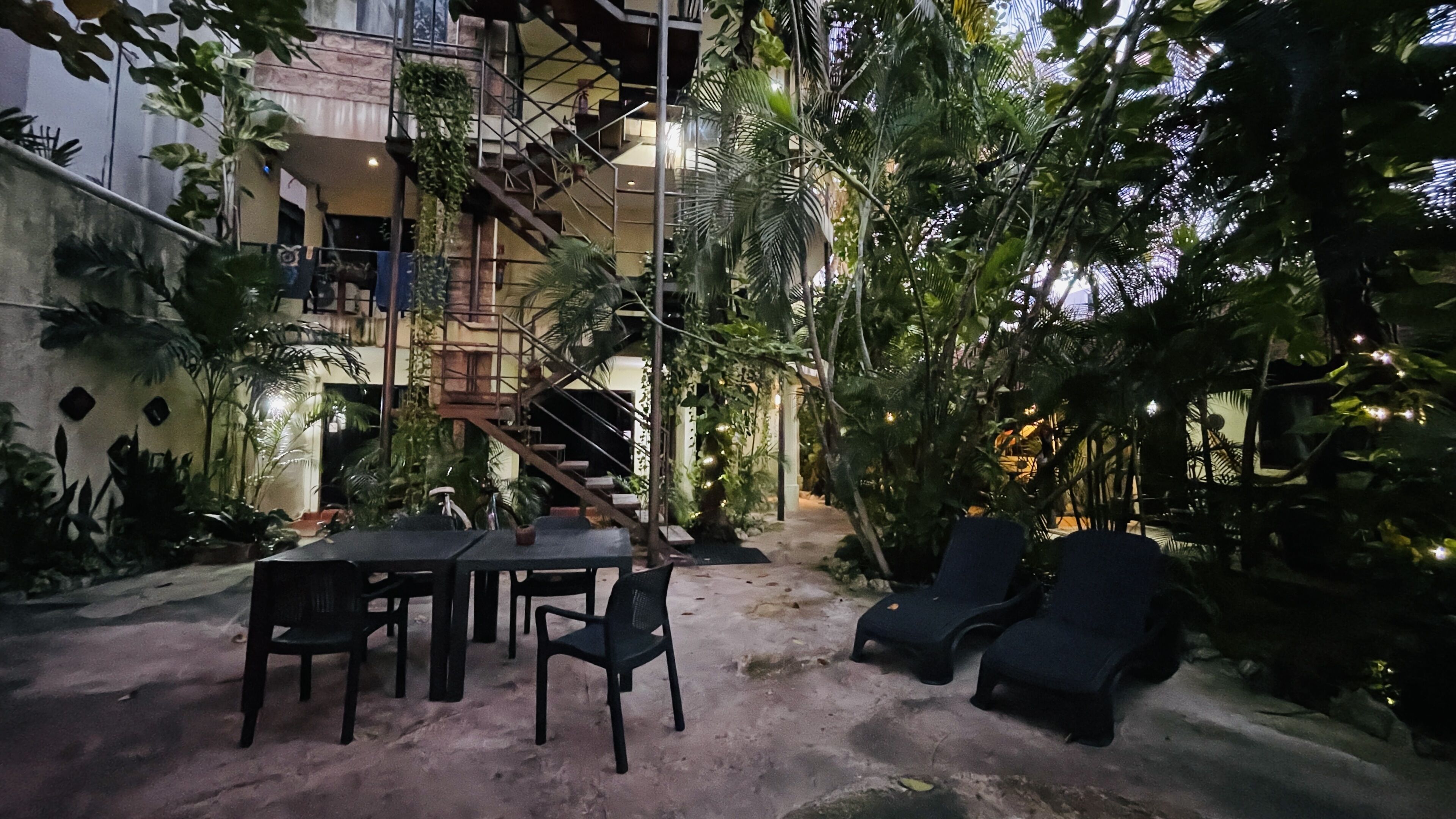 An outdoor seating area with a table and chairs, surrounded by lush greenery and a staircase leading to an upper level.