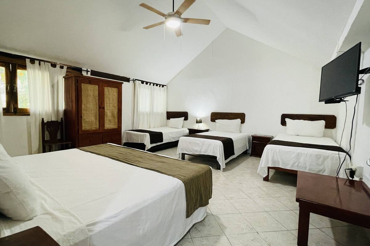 A hotel room with four single beds, a wooden wardrobe, a ceiling fan, a television mounted on the wall, and a window with curtains.