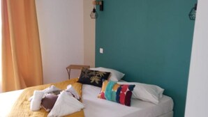 Ein Einzelbett mit gelber Decke, weißen Kissen und bunten Zierkissen. Dahinter eine türkis gefärbte Wand und eine weiße Klimaanlage an der Wand.