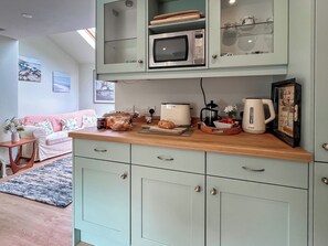 A kitchen with a microwave, toaster, coffee maker, and a fruit bowl. There is a sofa and a small table with a vase in the living area.