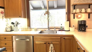 A kitchen with wooden cabinets, a stainless steel dishwasher, a window with a plant, a coffee maker, and a shelf with glasses. - Mount Snow Vermont - Ski - Snowboard - 6 miles (Wilmington)