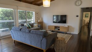A modern living room with a grey sofa, wooden flooring, a television, and a large window. - Relax in this boho coastal home walking distance to beautiful beach (Blairgowrie)