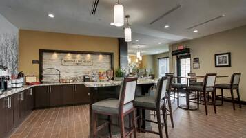 A hotel kitchen with a 'FREE DAILY BREAKFAST' sign, a buffet counter, and a dining area with chairs and tables.