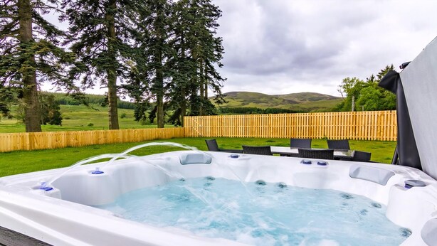 Ein Whirlpool mit einer Holzterrasse, umgeben von einem Holzzaun und Bäumen, mit einer Rasenfläche und Hügeln im Hintergrund.
