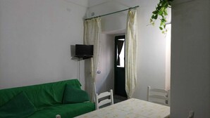 A small room with a green sofa, a table with chairs, a TV mounted on the wall, and a window with curtains. - Sandra's House in Meta di Sorrento (Meta)
