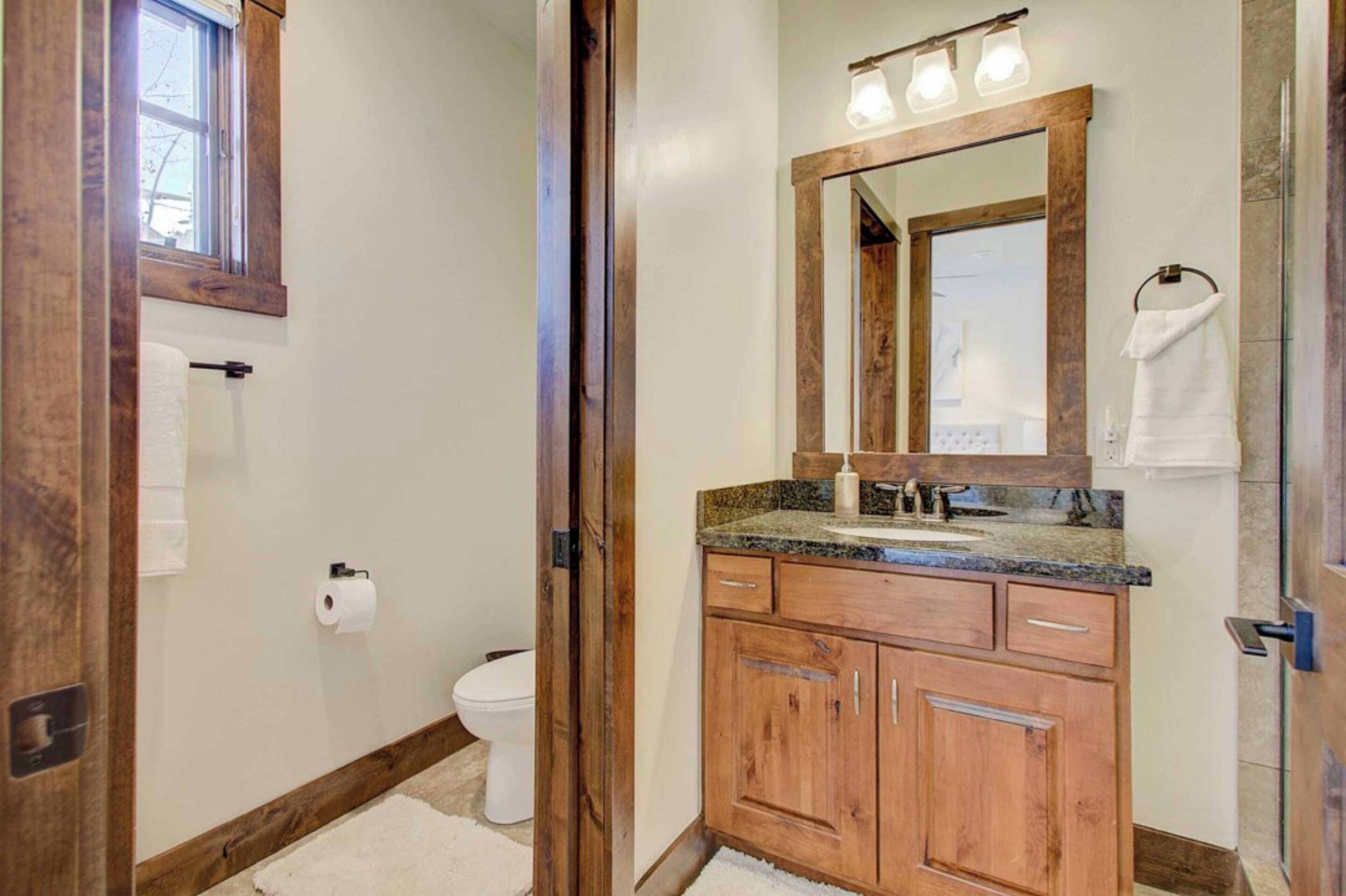 A bathroom with wooden cabinets, a granite countertop, a large mirror, and a window above the toilet.