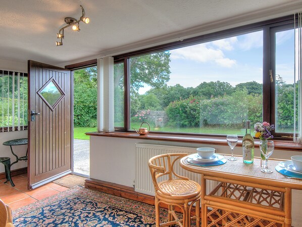 A dining area with a table set for two, a view of the garden through large windows, and a door leading outside.