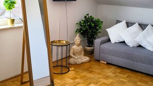 A living room with a wooden parquet floor, a grey sofa, a large mirror, a Buddha statue, and a potted plant.