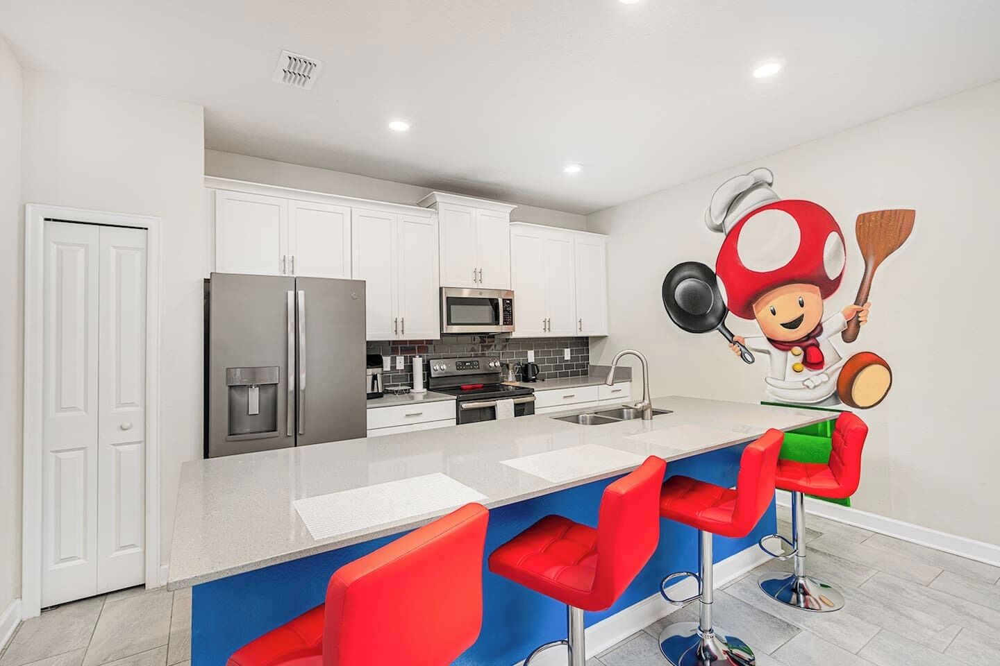 A modern kitchen with white cabinets, stainless steel appliances, a central island with red bar stools, and a cartoon character mural.