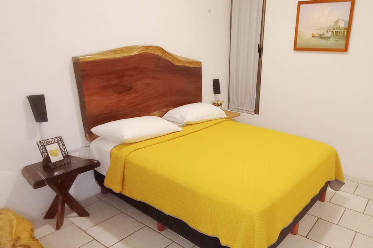 Un lit avec une tête de lit en bois, une couvre-lit jaune et des oreillers blancs. Il y a une table de chevet avec un cadre photo et une lampe. La pièce est dotée d’un sol carrelé blanc et d’une fenêtre avec un rideau blanc.