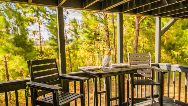 A wooden deck with a table set for two, surrounded by chairs, overlooking a forested area.
