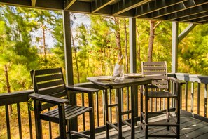 A wooden deck with a table set for two, surrounded by chairs, overlooking a forested area.