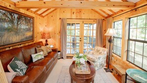 A cozy room with a wooden ceiling, a large window, a sofa, a coffee table, and a painting on the wall.