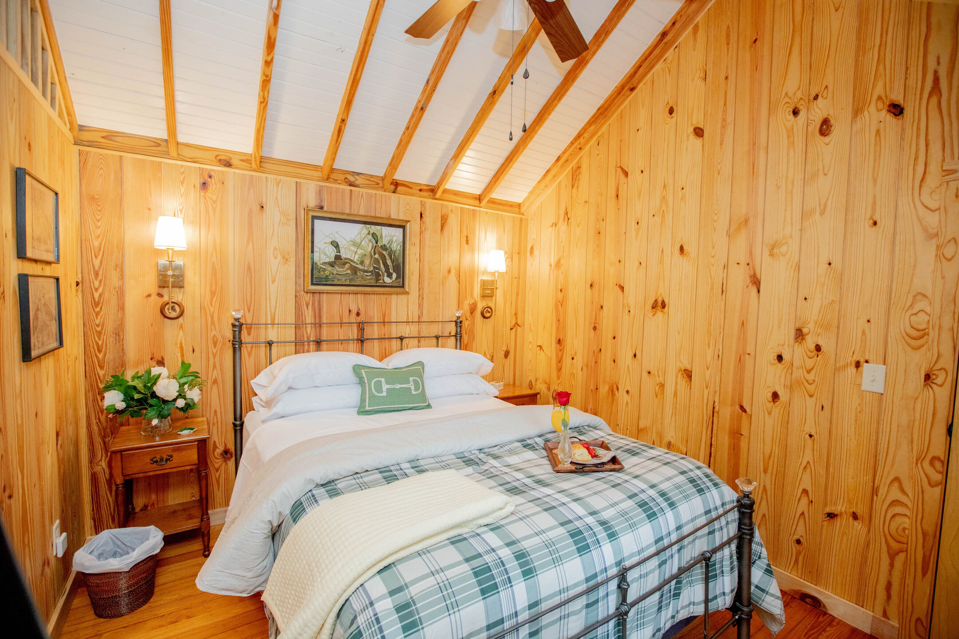A wooden-paneled bedroom with a bed, two bedside lamps, a nightstand, a wicker basket, and a framed picture on the wall.