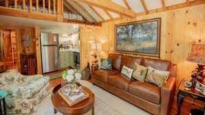 A cozy cabin interior with a wooden ceiling, a living room with a brown leather sofa, a floral armchair, and a dining area with a refrigerator.