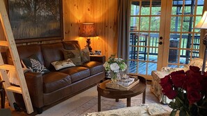 A cozy cabin interior with a wooden ceiling, a leather sofa, a coffee table, and a large window overlooking a deck.