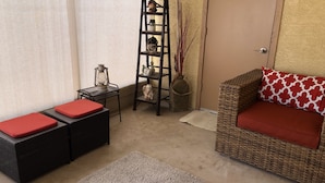 A cozy living area with a wicker sofa, red cushions, a small side table with a lantern, and a tall black shelving unit against a textured wall. - Peaceful and tranquil townhouse for seniors  Heated pool/Spa gym. covered patio (Mesa)
