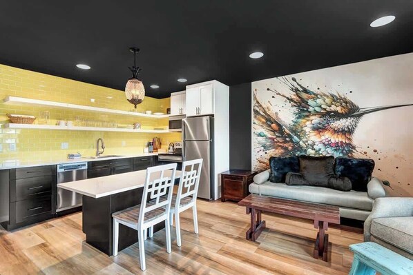 A modern kitchen with a central island, white chairs, a dining table, and a sofa with a colorful abstract painting on the wall. - Top Floor Riverfront Condo at the edge of Downtown (Bend)