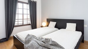 Et soveværelse med to senge, mørke gardiner, en senget lampe og et vindue med udsigt.