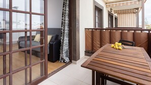 En balkon med et træbord og stole, en sofa samt en skydedør, der fører til et indre rum.