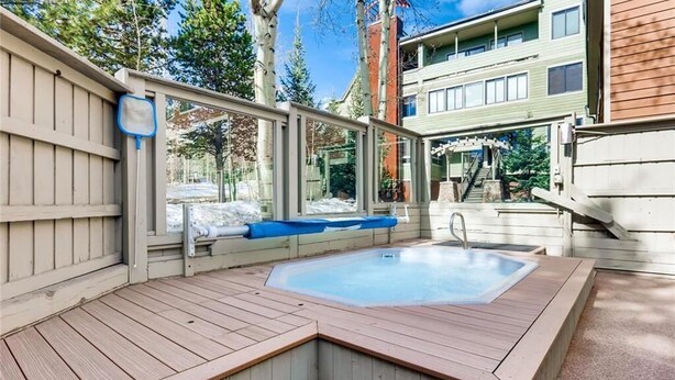 Ein Whirlpool mit Holzterrasse und Glasumrandung, umgeben von Bäumen und einem Gebäude im Hintergrund.