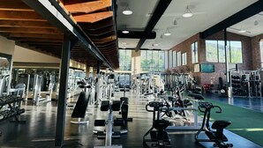 Uma academia espaçosa com diversos equipamentos de exercício, grandes janelas e um teto alto com vigas aparentes.