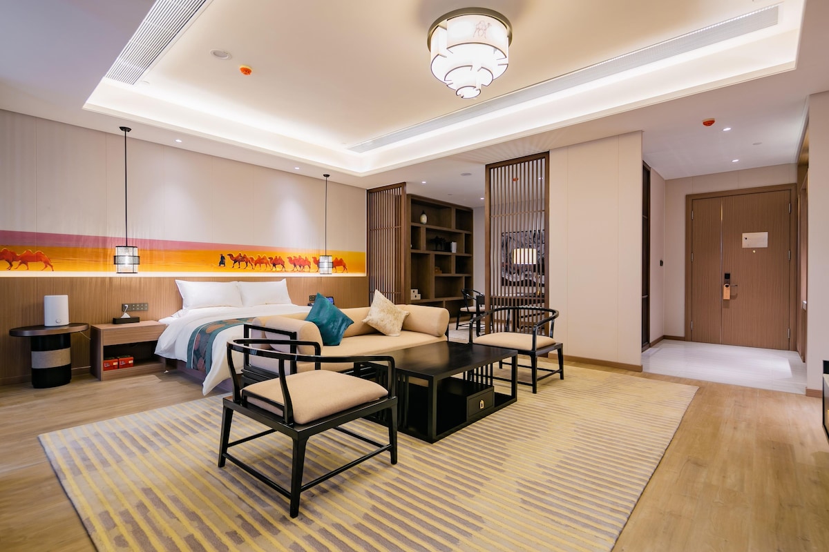 A hotel room with a bed, sofa, chairs, a coffee table, a nightstand, a lamp, a bookshelf, and a door.